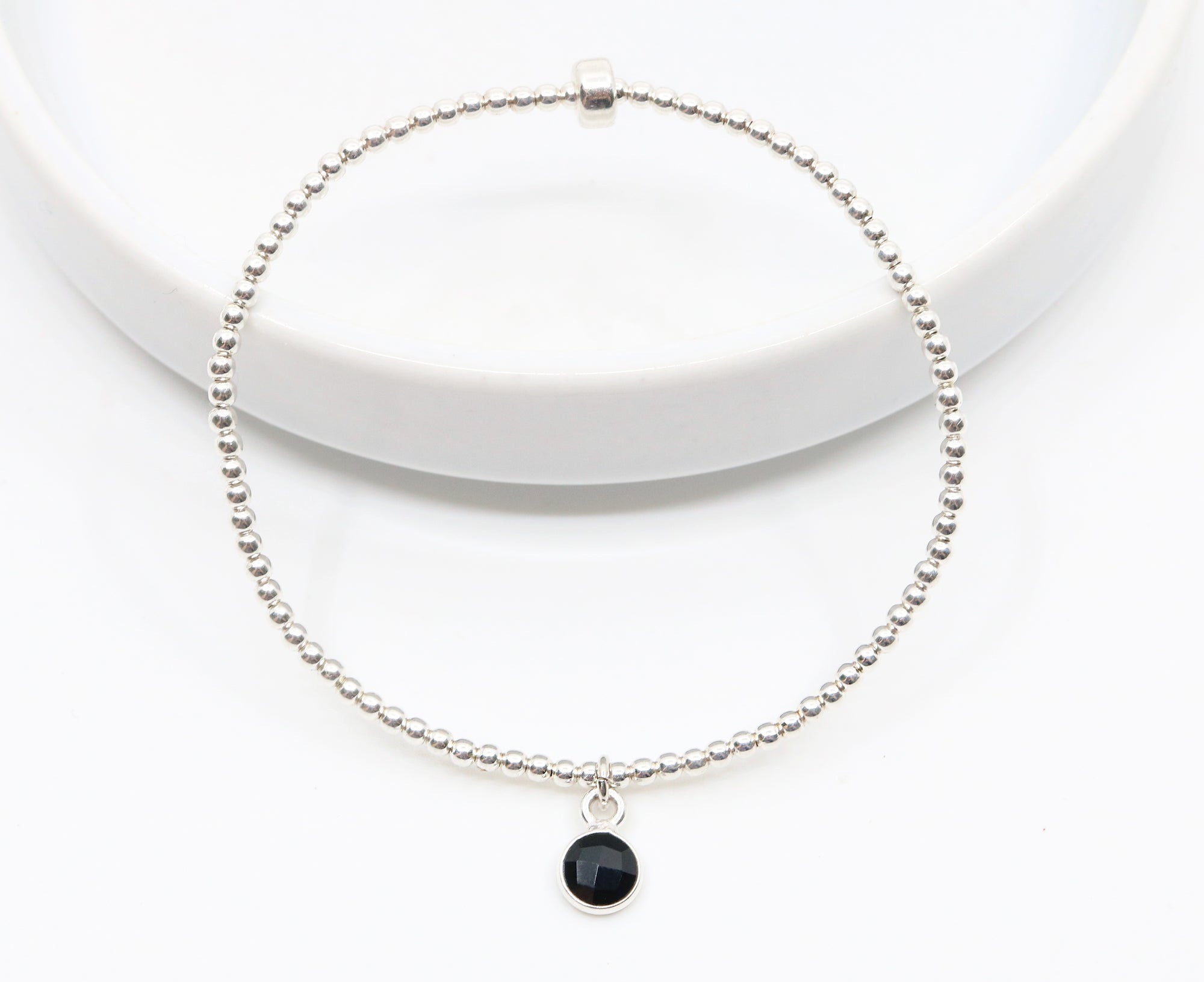 Small onyx gemstone circle pendant encased in sterling silver dangling from a sterling silver beaded stretch bracelet and displayed on white background