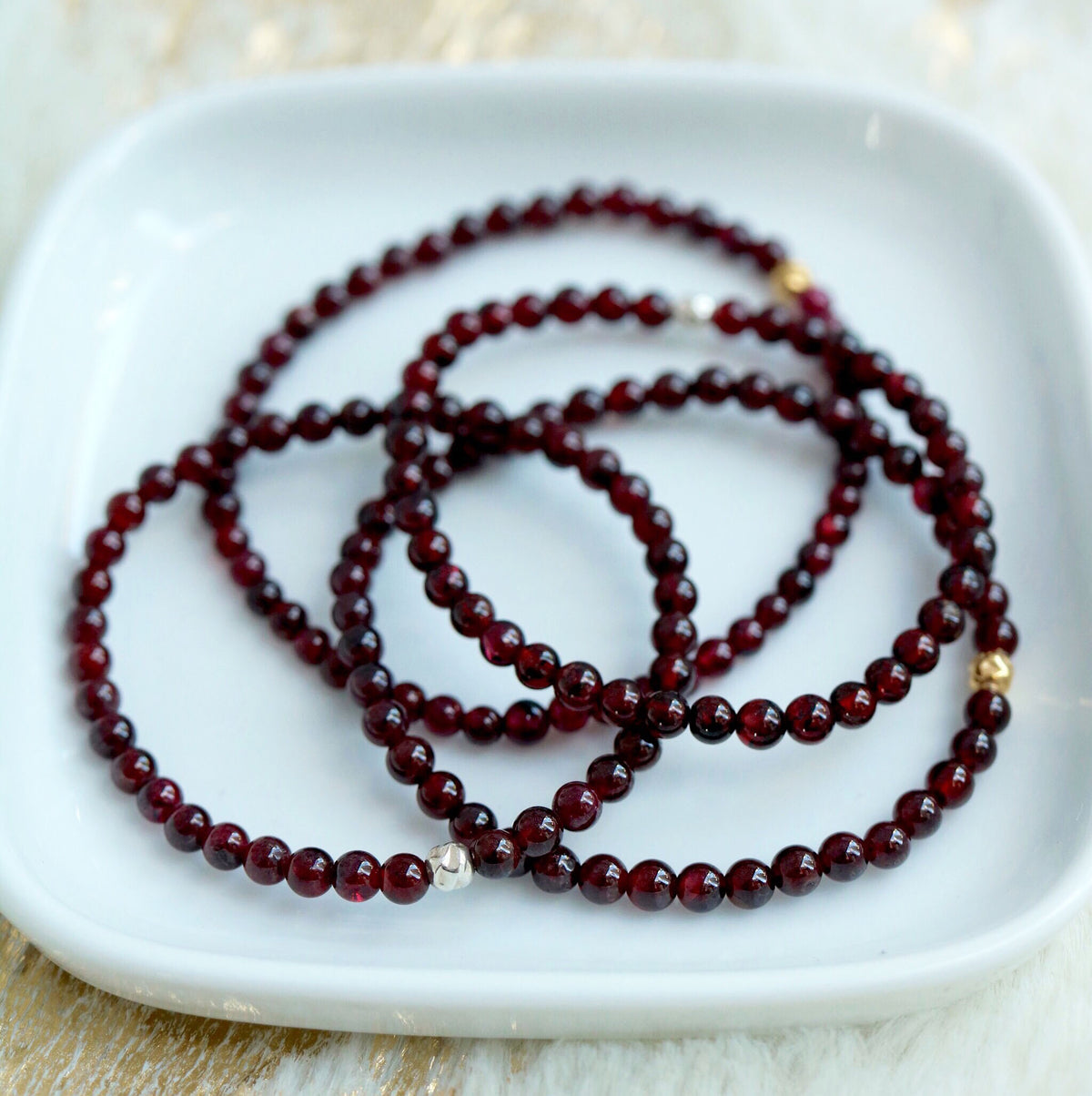 multiple garnet beaded stretch bracelets with either gold or silver hammered accent displayed in white ceramic dish
