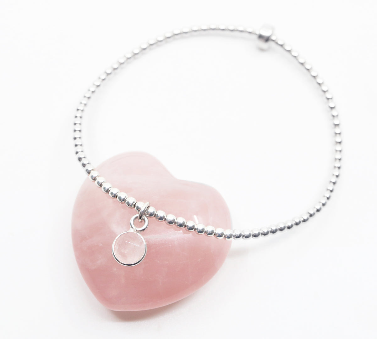 Small rose quartz circle pendant encased in sterling silver dangling from a sterling silver beaded stretch bracelet and displayed resting on heart shaped rose quartz crystal
